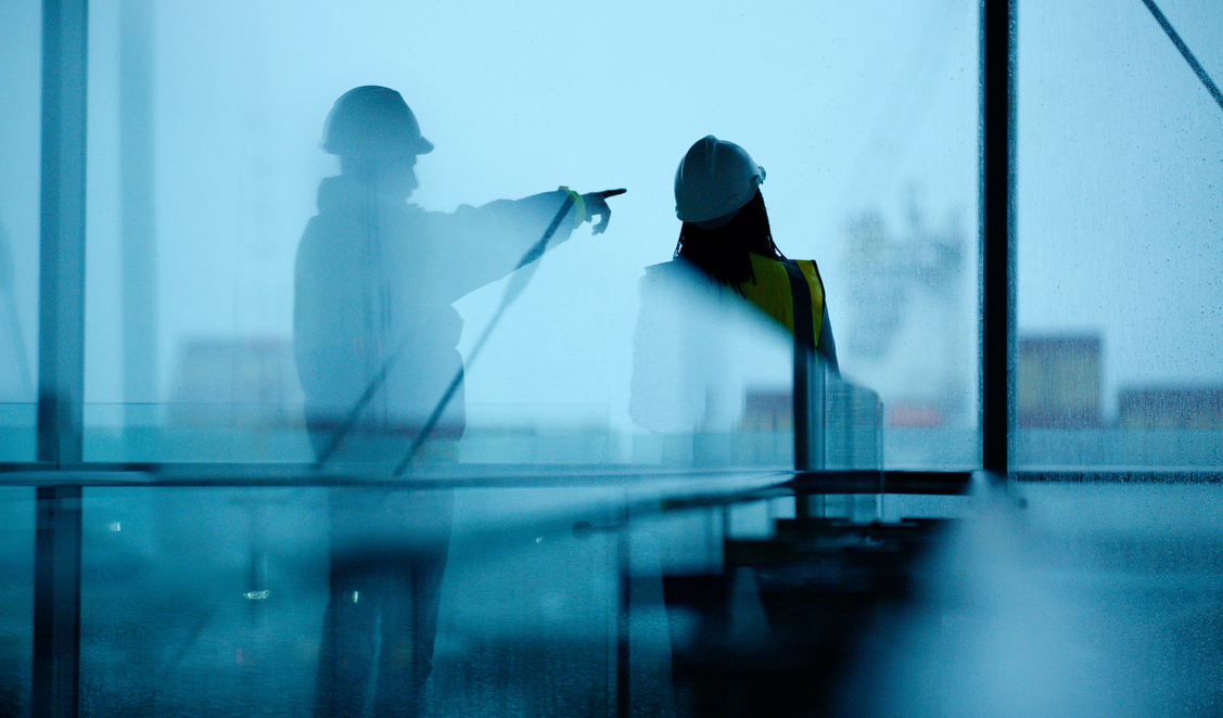 Two engineers in safety gear discussing plans through a glass wall, with one pointing forward.